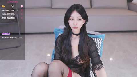 Media: Video of a young Asian woman with long black hair, wearing a sheer black top, black choker, and red stockings, sitting on a blue cushioned chair, in a modern living room.