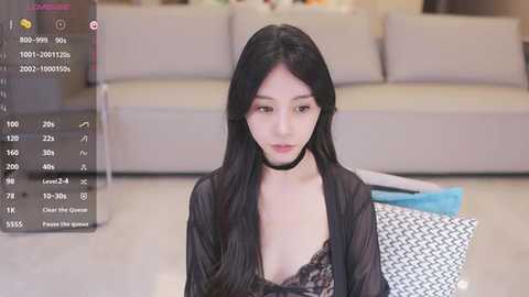 Media: Video of a young Asian woman with long black hair, wearing a black lace top and choker, sitting on a beige sofa in a modern living room.