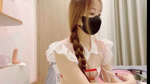 Media: Video of an East Asian woman with long, light brown hair in a braid, wearing a black face mask, white ruffled dress, and red lingerie. She's seated at a desk with a laptop and headphones. Background includes beige curtains and a wooden shelf.