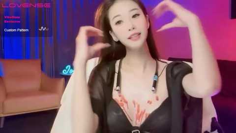 Media: Video of an East Asian woman with long black hair, wearing a black top with a large cleavage, performing a hand gesture in a modern living room with blue and pink lighting.