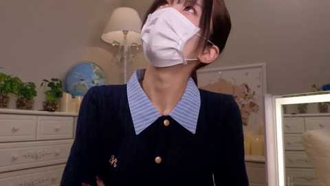 Media: Video of an Asian woman in a navy blue school uniform with a white mask, standing in a brightly lit bedroom with a dresser, globe, and framed artwork in the background.