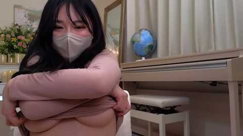 Media: Video of an East Asian woman with long black hair, wearing a pink top, face mask, and gloves, hugging herself in a beige room with a piano, floral decor, and a globe.