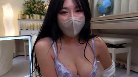 Media: Video of an Asian woman with long black hair, wearing a light blue lace bra, beige mask, and white robe, seated in a cozy, warmly lit living room with candles, a globe, and a floral arrangement in the background.