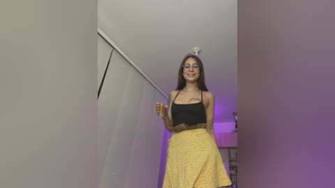 Media: Video of a smiling woman with long dark hair, wearing a black crop top and yellow floral skirt, standing in a narrow hallway with white walls and purple lighting.