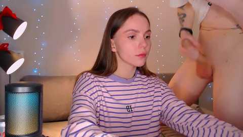 Media: A video of a young woman with straight brown hair wearing a striped sweater, looking towards a man with a visible erect penis, in a cozy room with a lamp and string lights.