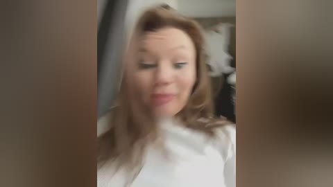 Media: Blurry video of a woman with long, light brown hair, wearing a white top, smiling, standing indoors with blurred background.