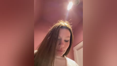 Media: Video of a young woman with long brown hair, wearing a white top, standing in a dimly lit room with reddish-brown walls. She appears to be looking down, with a soft, introspective expression.