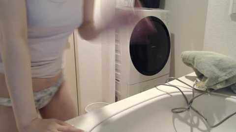 Media: A blurred video of a person in a white top and panties, standing beside a white washing machine in a laundry room.