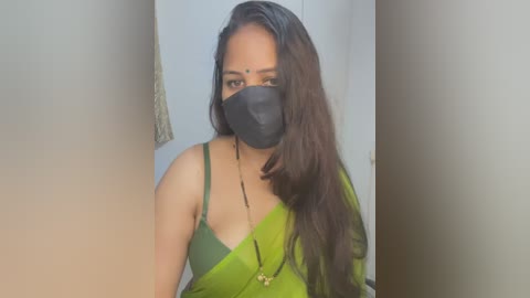 Media: Video of a South Asian woman with long, dark hair, wearing a green saree, black face mask, and black earrings. She stands indoors, against a plain white background.