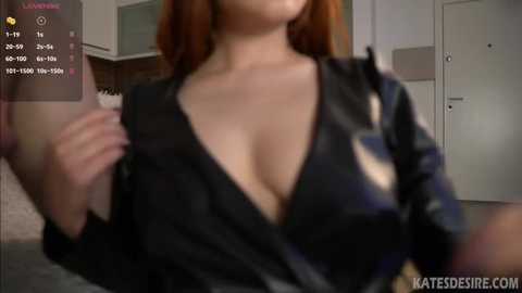 Media: A video of a woman with light skin and long, straight red hair, wearing a black dress with a deep V-neck, revealing cleavage, in a modern, brightly lit room with white walls and a door in the background.