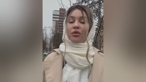 Media: Video of a Caucasian woman with light skin and brown hair, wearing a beige coat and white scarf, standing outdoors in a park with bare trees and high-rise buildings in the background.