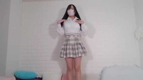 Media: A video of an Asian woman with long black hair, wearing a white shirt, plaid skirt, and mask, standing against a white brick wall in a room with white furniture.