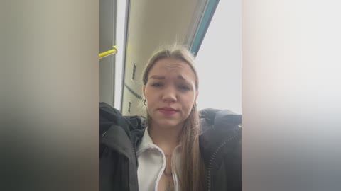 Media: Video of a young woman with straight, light brown hair, wearing a black jacket and white top, sitting in a bus. She has a neutral expression. Background includes a yellow pole and a window.