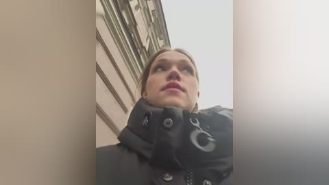 Media: A video of a Caucasian woman with blonde hair, wearing a black puffer jacket, gazing upwards against a beige building.