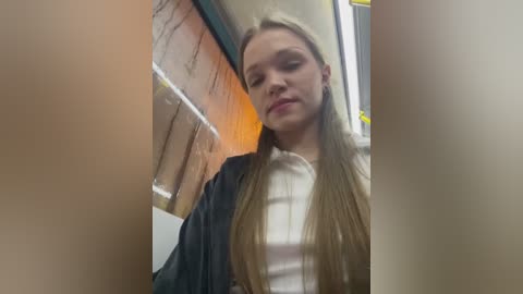 Media: Video of a young Caucasian woman with long, straight, light brown hair, wearing a white top and dark jacket, standing in a subway car with brick walls and yellow lighting.