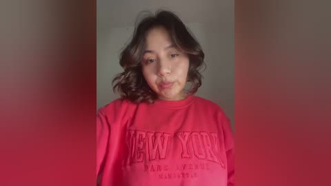 Media: Video of an Asian woman with shoulder-length, wavy brown hair, wearing a red New York Yankees sweatshirt, against a blurred red background.