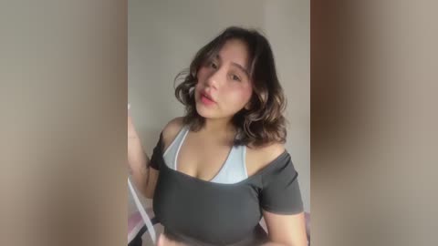 Media: Video of a young Asian woman with shoulder-length wavy hair, wearing a black off-shoulder top, posing indoors against a plain beige wall.