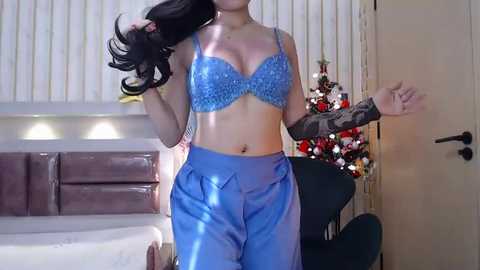 Media: Video of a woman in a blue sequined bra and high-waisted pants, holding her long black hair, standing beside a Christmas tree in a modern living room.