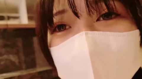 Media: Video of an Asian woman with fair skin, brown hair, and brown eyes, wearing a white surgical mask. Background features blurred kitchen cabinets and a stove.