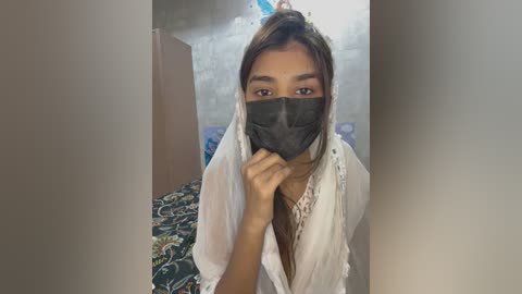 Media: Video of a young South Asian woman with light brown skin, wearing a white saree and dark face mask, standing in a dimly lit room with a floral-patterned bedspread and white wall.