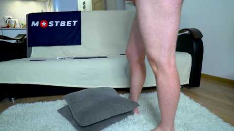 Media: Video of a nude, fair-skinned man standing on a white rug in a modern living room, placing a grey cushion on a beige couch. A \"MOSTBET\" banner hangs behind the couch.