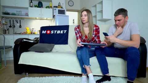 Media: Video of a blonde woman in a red plaid shirt, blue jeans, and white socks on a beige sofa, watching a man in a grey t-shirt and blue jeans play a video game. Modern kitchen background.