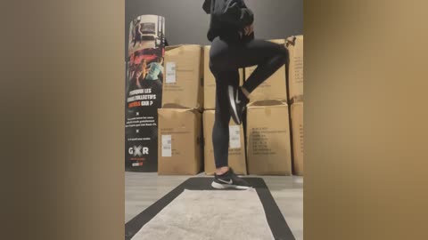 Media: Video of a person in black leggings and sneakers performing a kick on a mat in a warehouse with stacked cardboard boxes and a poster in the background.