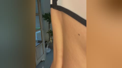 Media: Video of a close-up of a woman's bare torso, showing a mole on her right side, standing near a potted plant in a brightly lit room with white walls.