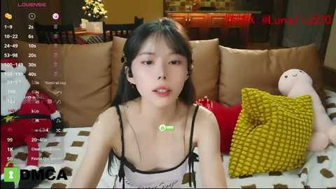 Media: A video of an East Asian woman with long black hair, wearing a white camisole, lying on a bed with a yellow pillow. The background shows a cozy room with brown cushions and a red Christmas tree.