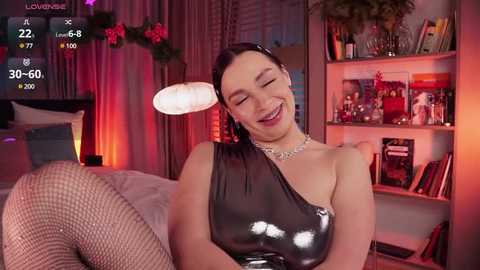 Media: Video of a smiling, light-skinned woman with dark hair, wearing a shiny, black one-shoulder latex dress and fishnet stockings, in a cozy, dimly lit bedroom with shelves of books and a bed.