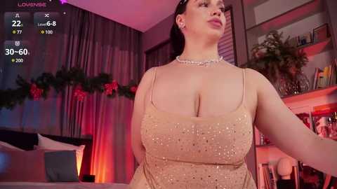 Media: Video of a curvy, fair-skinned woman with dark hair, wearing a glittery gold dress and pearl necklace, in a cozy bedroom with holiday decorations, red curtains, and a bed.