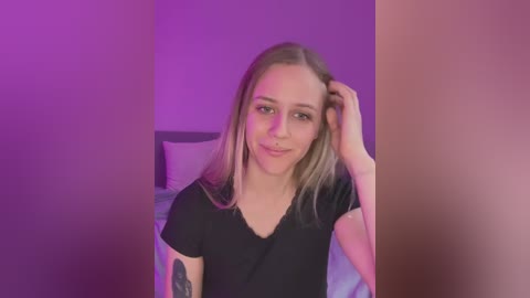 Media: Video of a young Caucasian woman with blonde hair, wearing a black t-shirt, standing in a bedroom with purple lighting. She has a tattoo on her left arm and is touching her hair with her right hand.