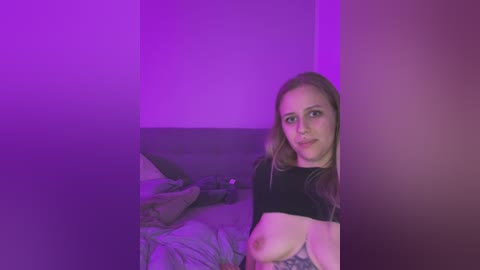 Media: A video of a young Caucasian woman with long blonde hair, wearing a black crop top, exposing her small breasts and nipples. The background features a dimly lit bedroom with purple lighting, unmade bed, and a pillow.