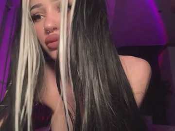 Media: Video of a young woman with long, straight, black and blonde hair, wearing a black top, with a nose ring and slightly parted lips, in a dimly lit room with purple lighting.