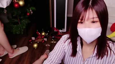 Media: Video of an Asian woman with long dark hair, wearing a white face mask, seated on a wooden floor, surrounded by festive Christmas decorations and a person in the background.