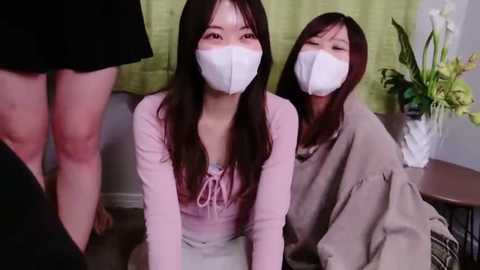 Media: Video of two East Asian women wearing white masks, pink tops, kneeling on beige carpet, in a room with green curtains and a vase of white flowers.