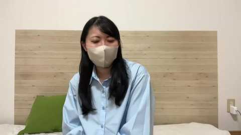 Media: A video of an Asian woman with long black hair, wearing a light blue shirt and beige surgical mask, sitting on a bed with a light wooden headboard, green pillow, and white sheets in a simple, clean room.