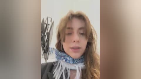 Media: Video of a young woman with long, wavy, light brown hair, wearing a blue and white patterned scarf and a grey jacket, standing in front of a blurred, beige background.