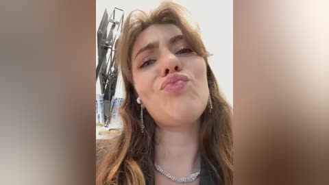 Media: Video of a young woman with fair skin, long brown hair, and a slightly pouty expression, blowing a kiss. She wears a silver necklace and has a backdrop of a metallic sculpture and a white wall.