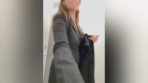 Media: Video of a woman in a gray blazer, holding a black jacket, taken indoors with blurred background.