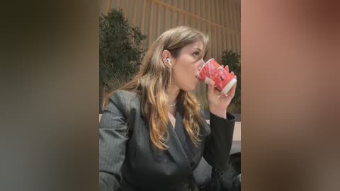 Media: Video of a woman with long, wavy, light brown hair, wearing a black blazer, drinking from a red cup, against a blurred background with a wooden fence and greenery.