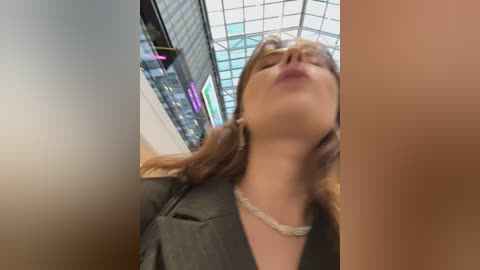 Media: A blurry video of a woman with light brown hair and fair skin, wearing a black jacket, standing in an indoor setting with a glass ceiling and modern architecture.