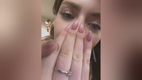 Media: A close-up video of a fair-skinned woman with brown hair, wearing pink glittery nail polish and a silver ring, covering her mouth with her hand. Background shows blurred decor.