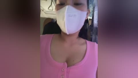 Media: Video of an Asian woman wearing a pink blouse and white surgical mask indoors, with blurred faces in the foreground.