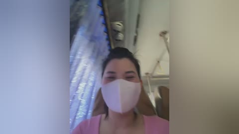 Media: A blurry video of an Asian woman with dark hair, wearing a pink top, white mask, and clear plastic shield, indoors, possibly in a medical setting.