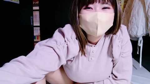 Media: Video of an East Asian woman with straight, shoulder-length brown hair, wearing a beige surgical mask, lavender blouse, and a white apron. She has light skin and a pleasant expression. Background features a bookshelf and a hanging garment.