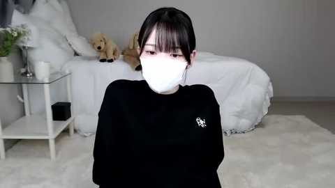 Media: Video of an East Asian woman with short black hair, wearing a black sweater and white mask, standing in a minimalist bedroom with a white bed, teddy bear, and glass table.