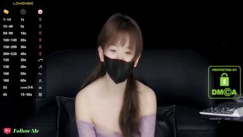 Media: A video of an East Asian woman with long, straight brown hair, wearing a black face mask and a light purple off-shoulder top, sitting on a black leather couch. The background shows a dimly lit room with a TV screen displaying live stream statistics.