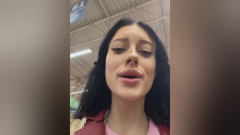 Media: A video shows a young woman with long black hair, pale skin, and a pink shirt, standing in a grocery store with a metallic ceiling and various shelves in the background.