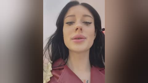 Media: Video of a young woman with pale skin, long black hair, and light makeup. She has a septum piercing, wearing a red jacket with a ruffled collar. Background is blurry, focusing on her face.